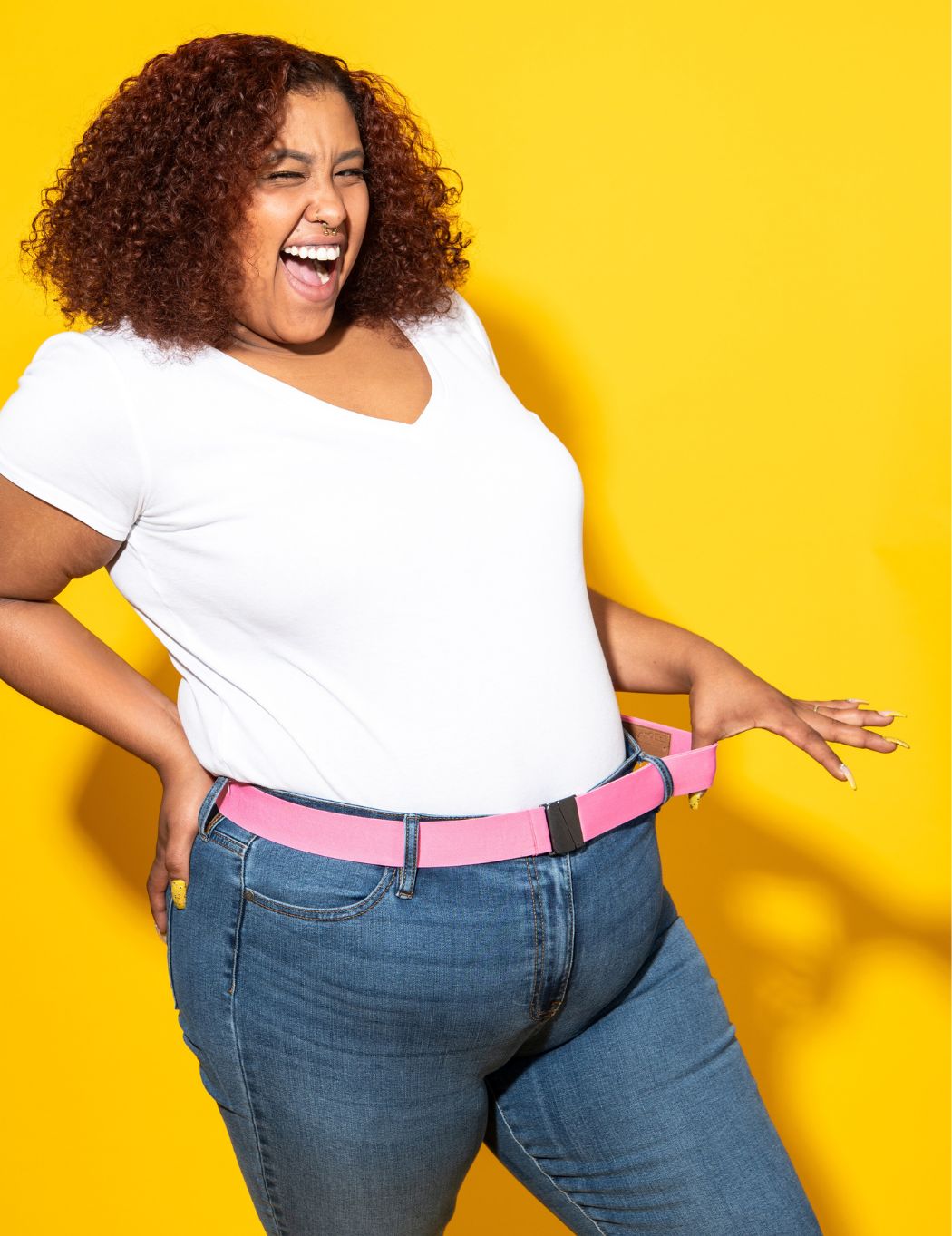 Image is a plus size woman wearing a white t-shirt and jeans with a pink Unbelts Intrepid stretch belt. She is stretching the belt elastic, winking and smiling a huge smile. Background is bright yellow.